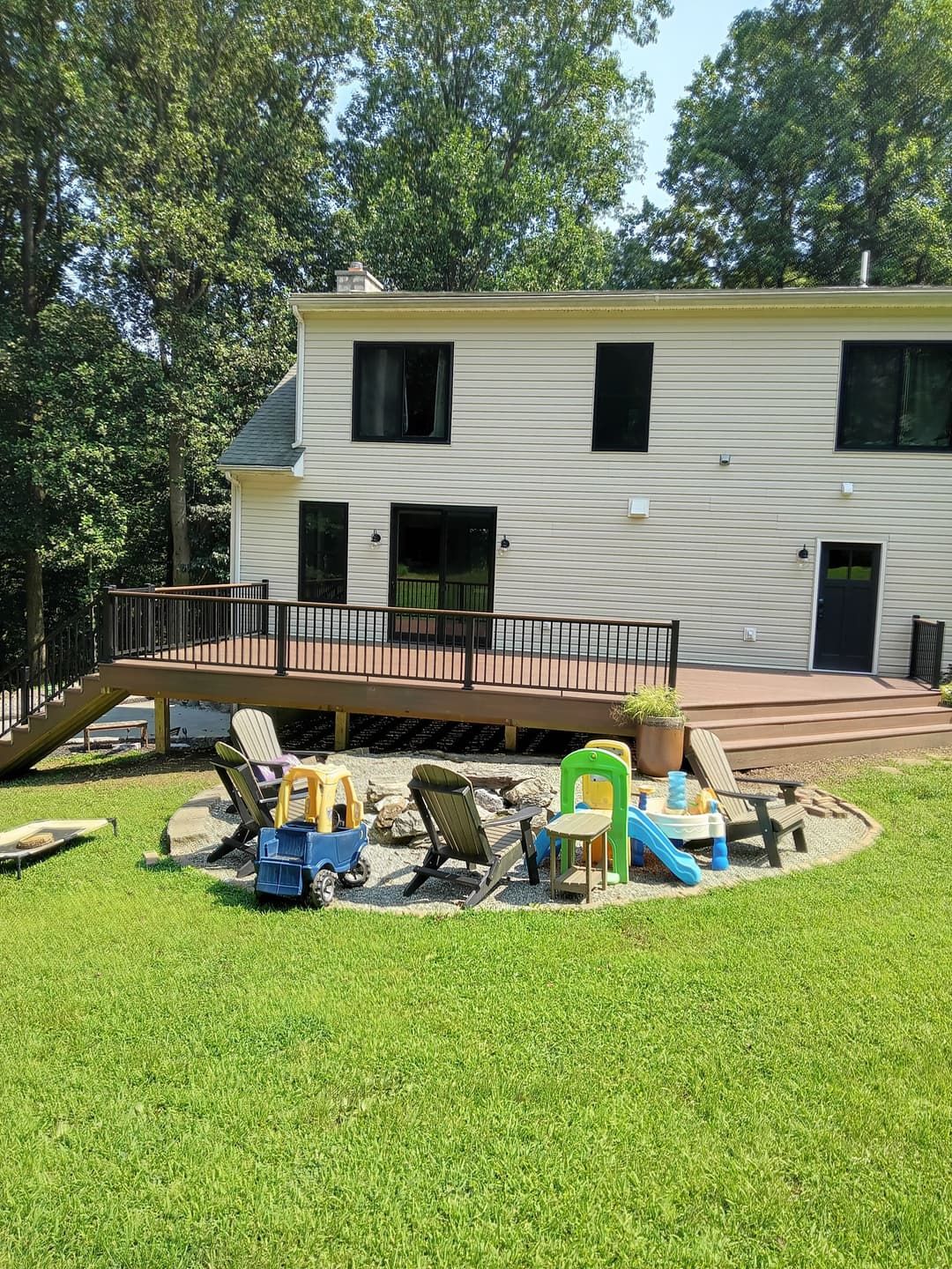 Backyard with a deck, fire pit, and children's toys on green grass, two-story house in the background.