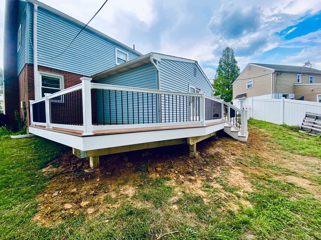 Wooden deck attached to a light blue house on a grassy hill. White railing, brown deck. Overcast sky.