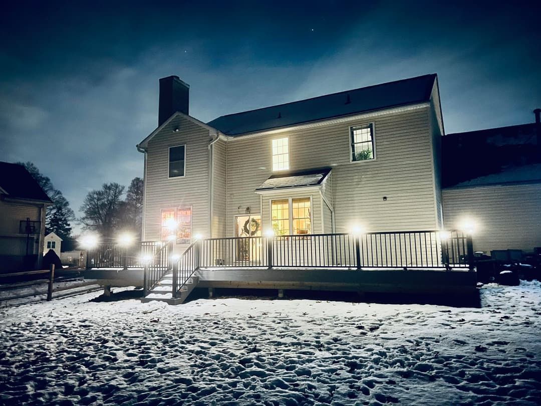Snowy night scene of a two-story house with deck, lit with outdoor lights. Window lights glow.