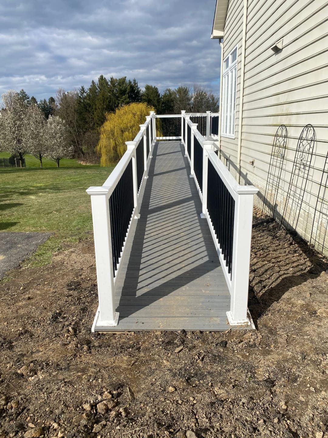 Ramp with gray deck and black/white railing leading to a house. Brown dirt surrounds the ramp.