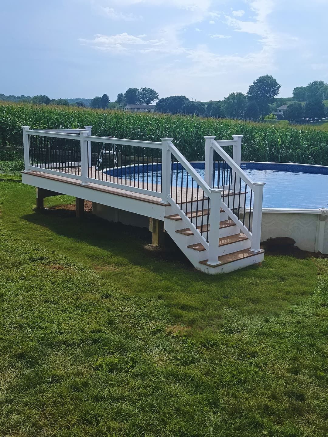 Deck with white railings and steps beside an above-ground pool, with a green lawn and fields in the background.