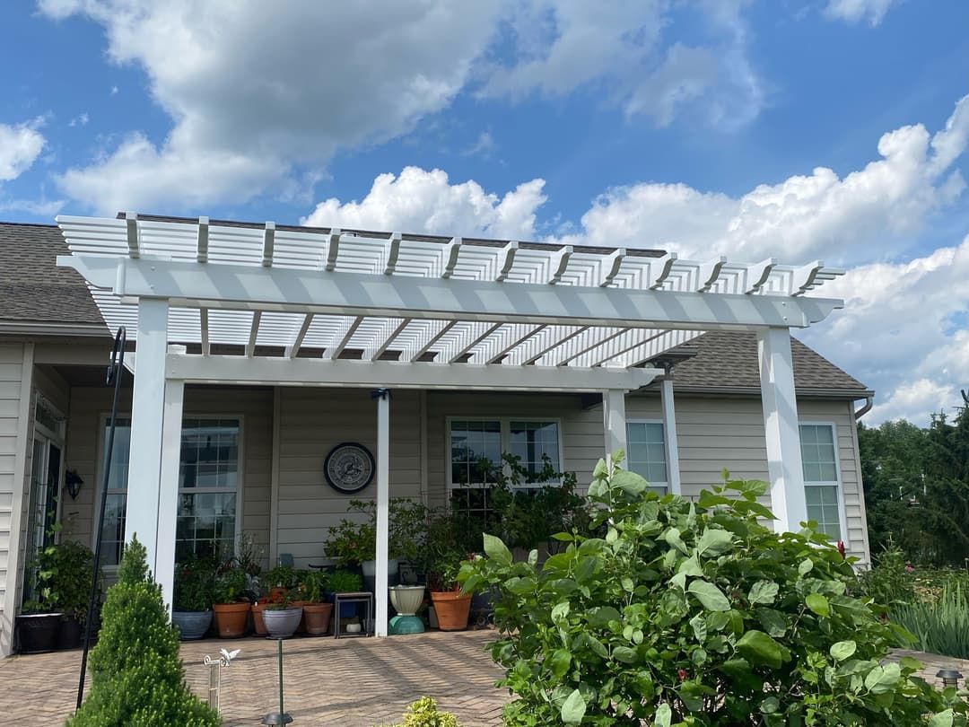 White pergola over a patio, with potted plants and a house in the background under a blue, cloudy sky.