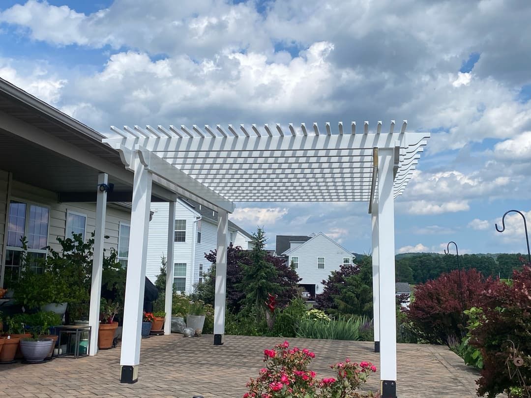 White pergola over a brick patio, with potted plants and flowering bushes. Blue sky with clouds.