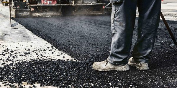 A man is standing in front of a truck spreading asphalt on a road.