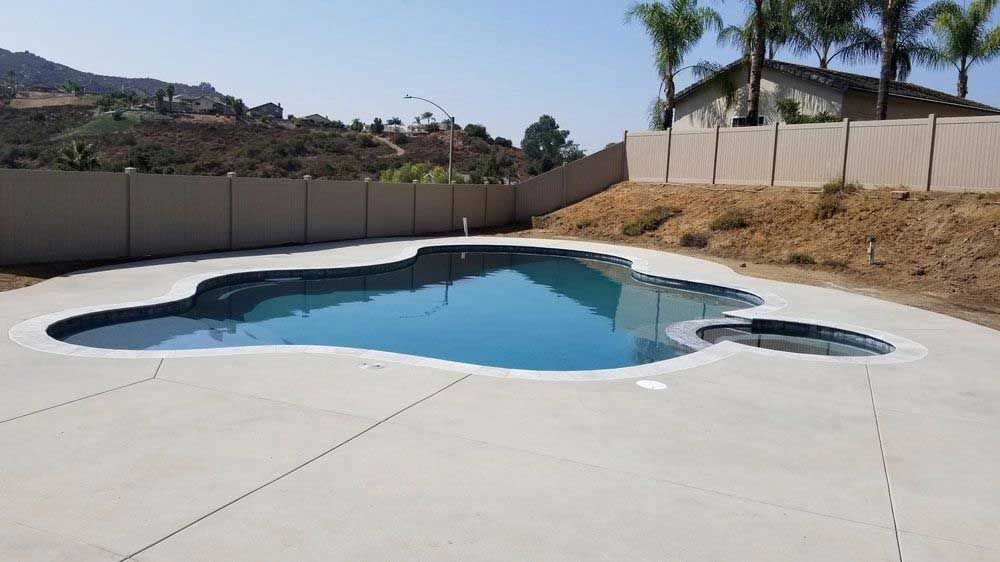 Empty backyard swimming pool with a curved blue-water section and white concrete deck, surrounded by a fence and hills.