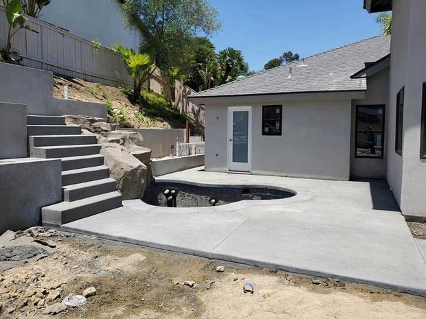 Concrete backyard patio with a small round pool, stairs, and a white house under a bright sky