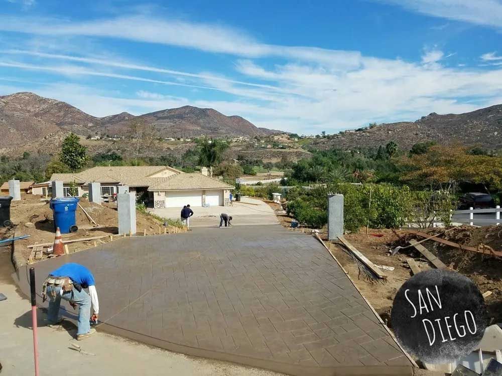Workers paving a concrete driveway in a hilly landscape, with mountains and a San Diego sign in view
