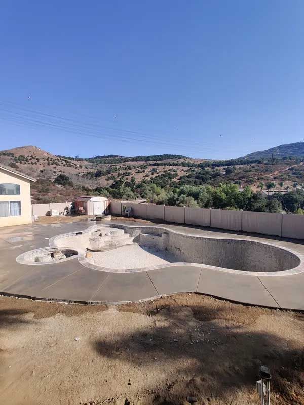 Outdoor empty swimming pool with a curved concrete deck, set against a hillside and blue sky.
