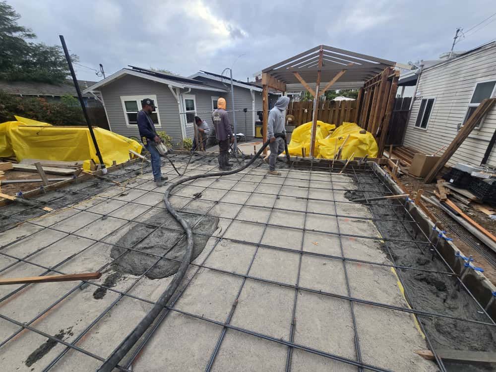 Concrete slab construction site with workers, rebar grid, and yellow tarps beside a house