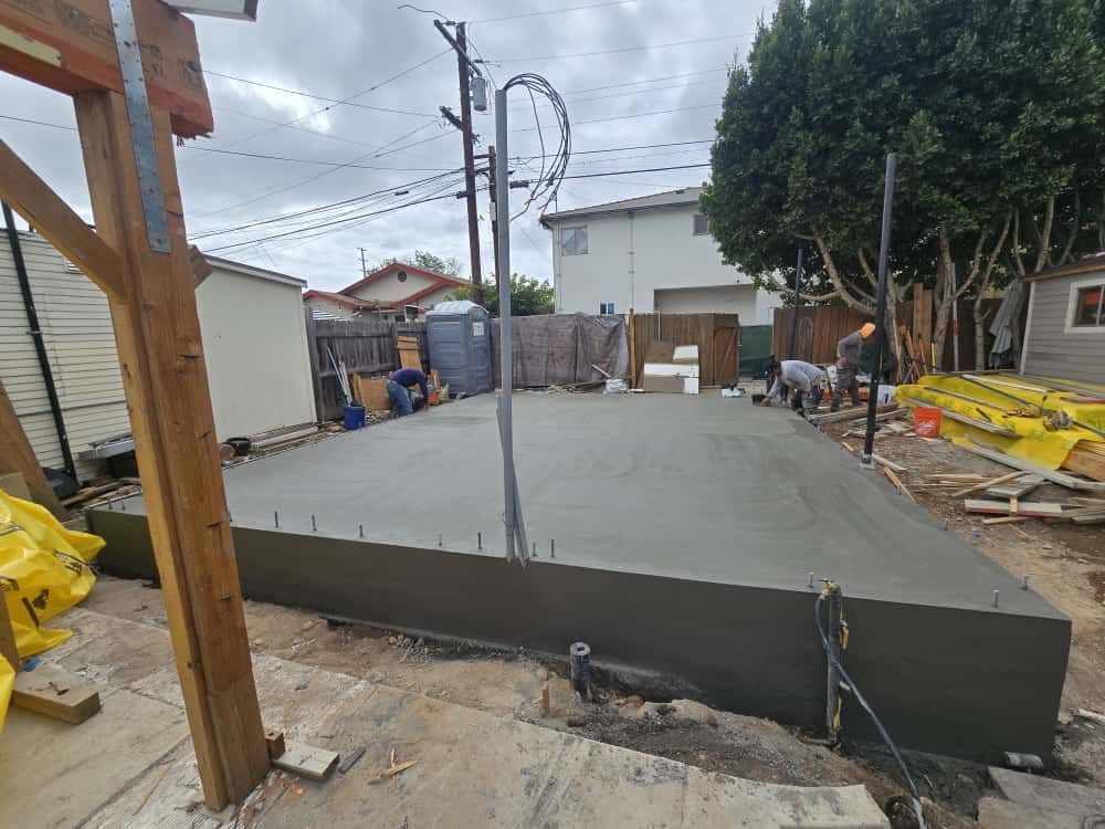 Concrete slab under construction in a backyard, with wooden forms, utility poles, and neighboring houses visible.