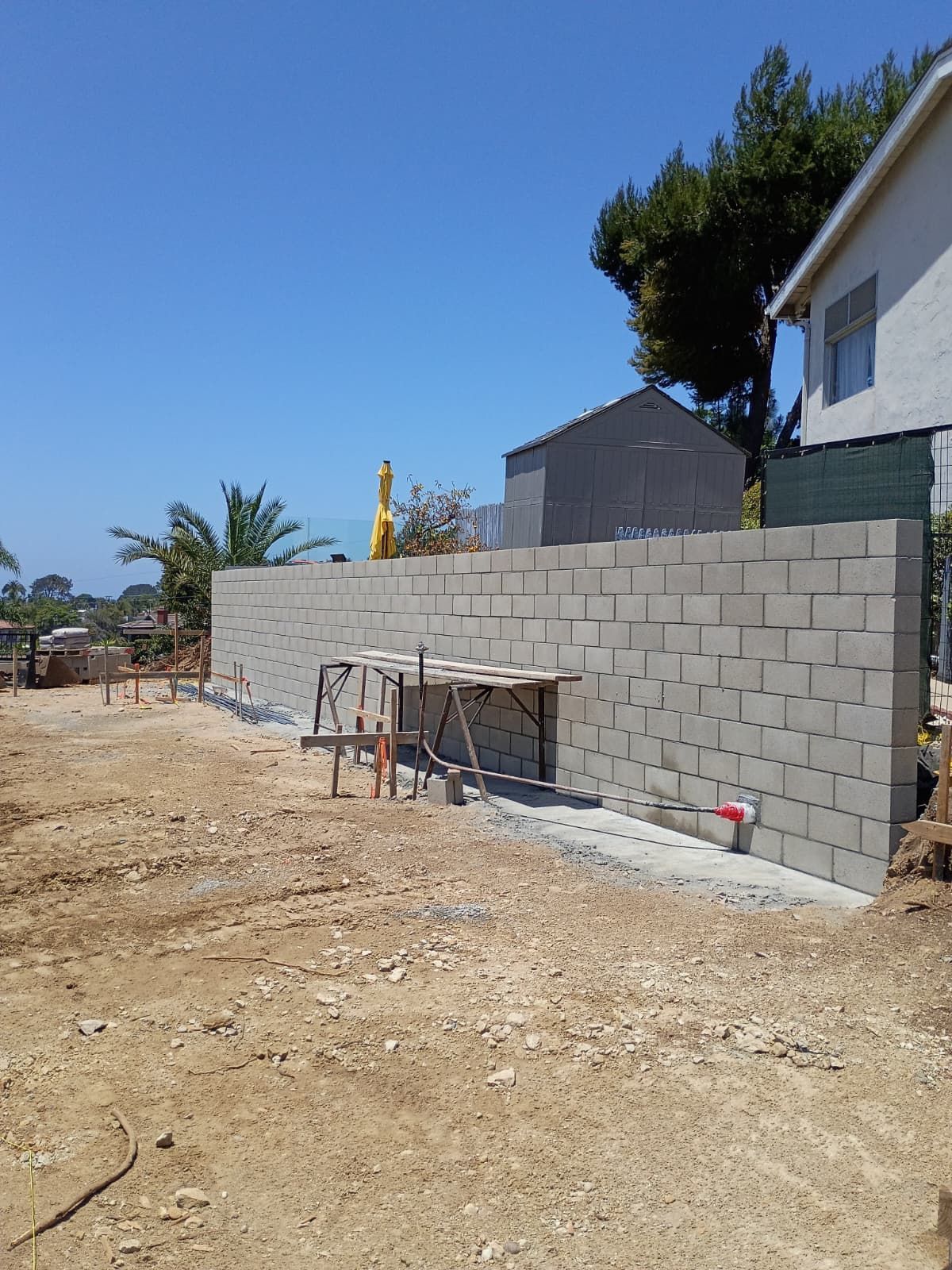 Partially built cinder block wall beside a dirt lot and house under a clear blue sky