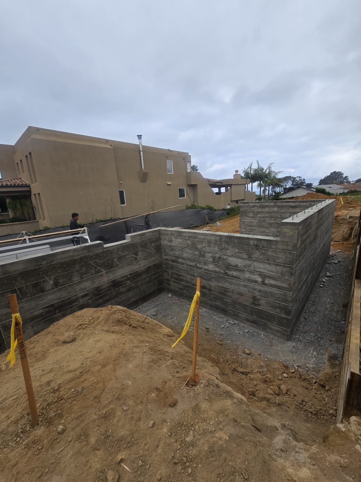 Construction site with concrete retaining walls, dirt foreground, and houses under a cloudy sky