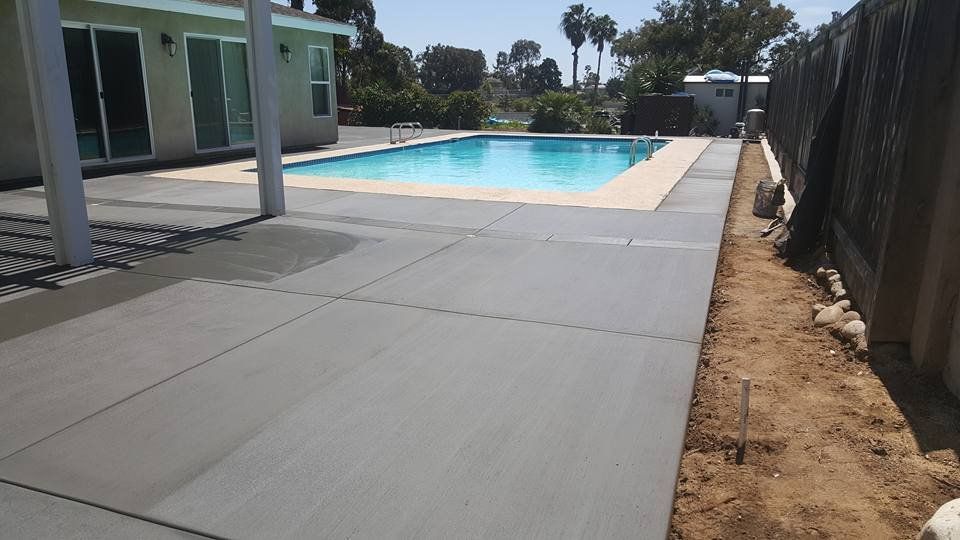 Concrete pool deck beside a swimming pool, with a fence and patio area in bright sunlight.