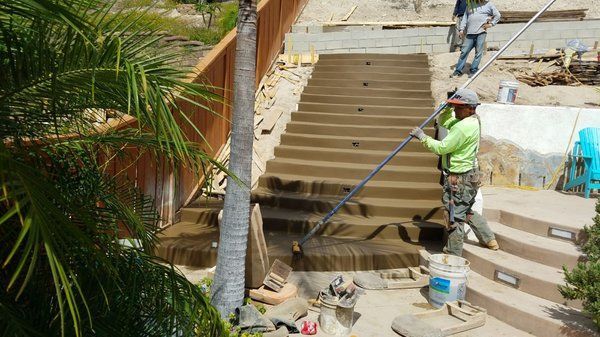 Construction workers pouring concrete on outdoor steps beside a palm-lined path