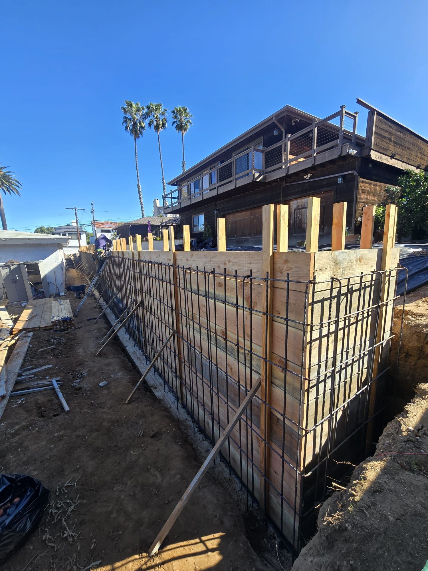 Outdoor construction site with rebar wall forms beside a wooden house and palm trees under a clear blue sky