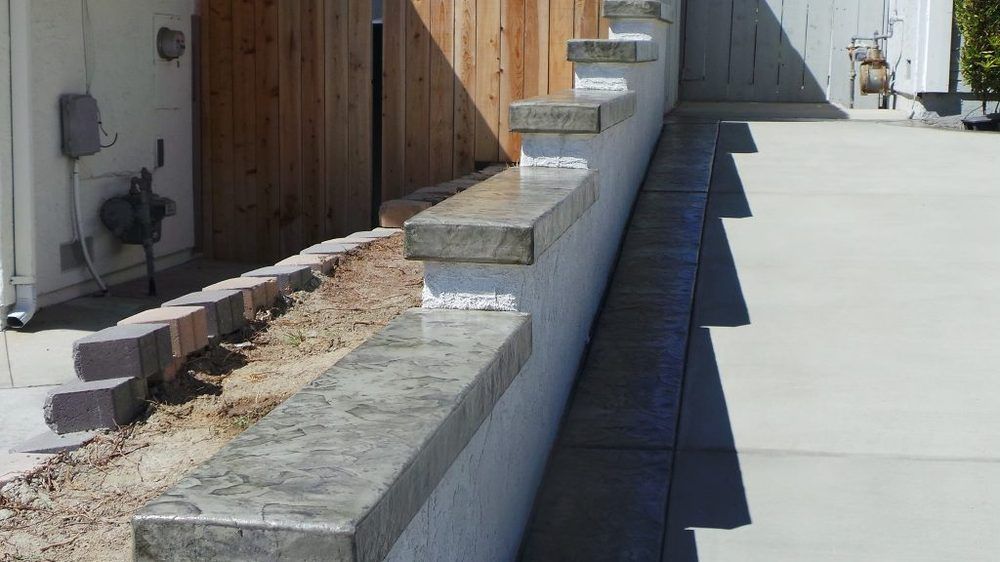 Concrete retaining wall along a narrow walkway beside a wooden building in bright sunlight