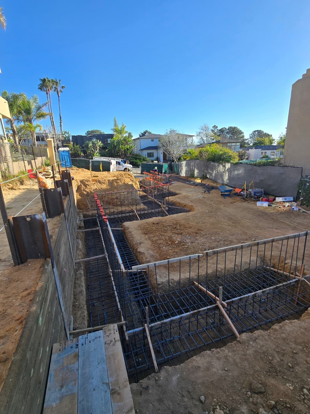 Construction site with rebar foundation forms, dirt excavation, and a house under construction in the background