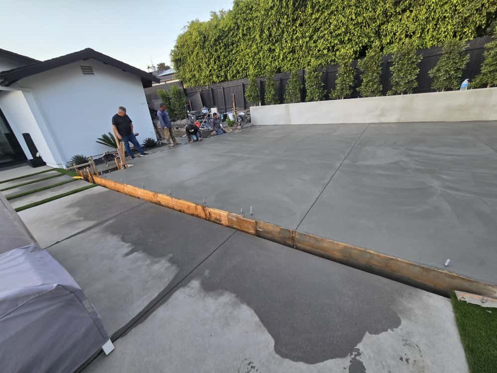 Backyard patio with a low wooden ramp, gray paving, and people near a white house and hedge.