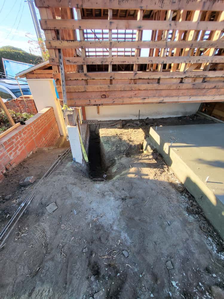 Construction area beneath a raised wooden deck with dirt ground, exposed beams, and a brick wall