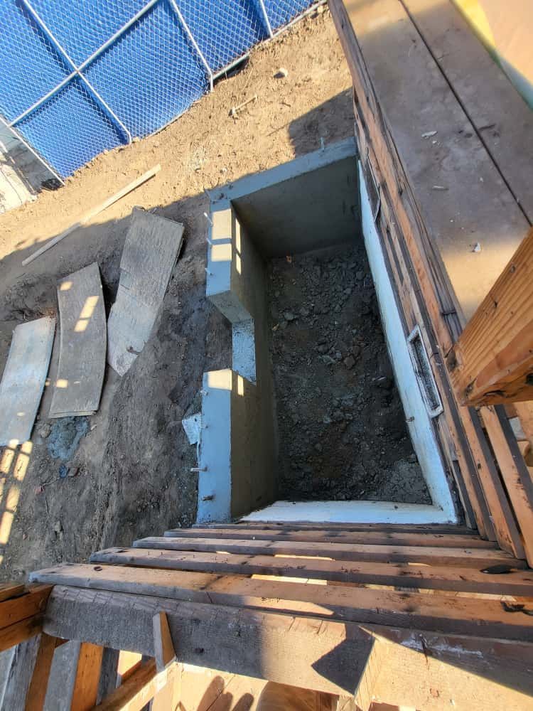 Open rectangular concrete pit beside wooden deck, with dirt and gravel inside
