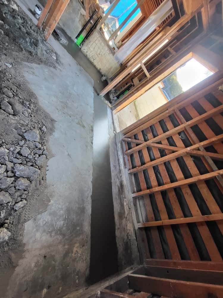 Narrow unfinished alley beside a wooden fence and concrete wall, with rough ground and construction debris