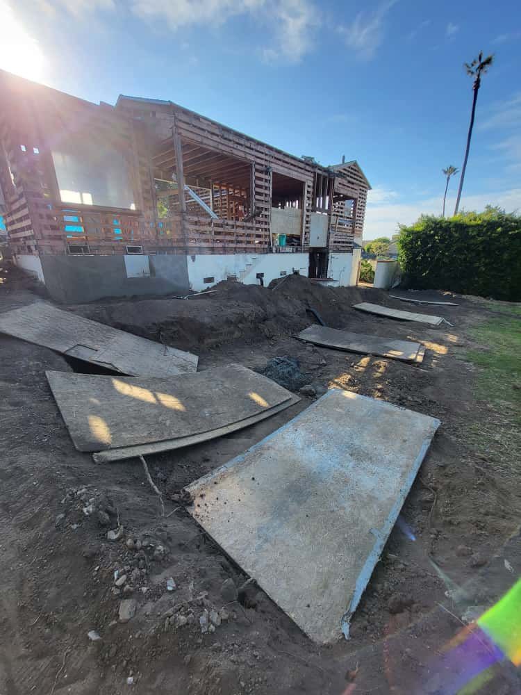 Partially demolished brick house with debris and concrete slabs scattered in a dirt yard, sunlit outdoors