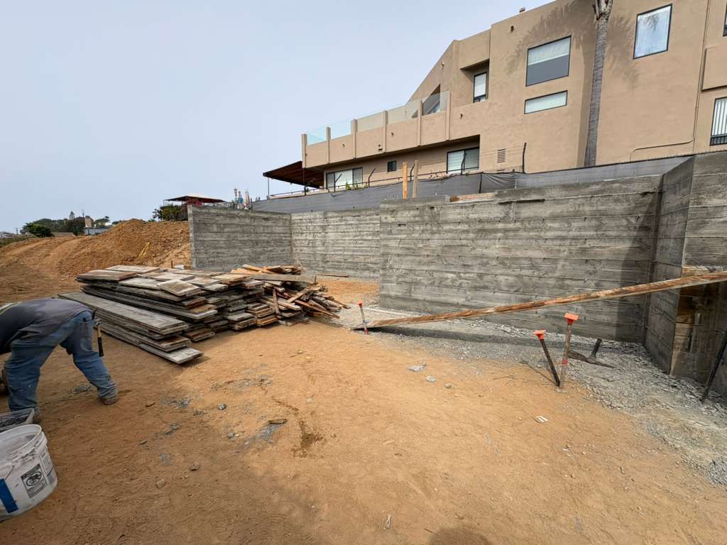 Construction site with concrete wall, stacked materials, and a worker kneeling on dirt near a building