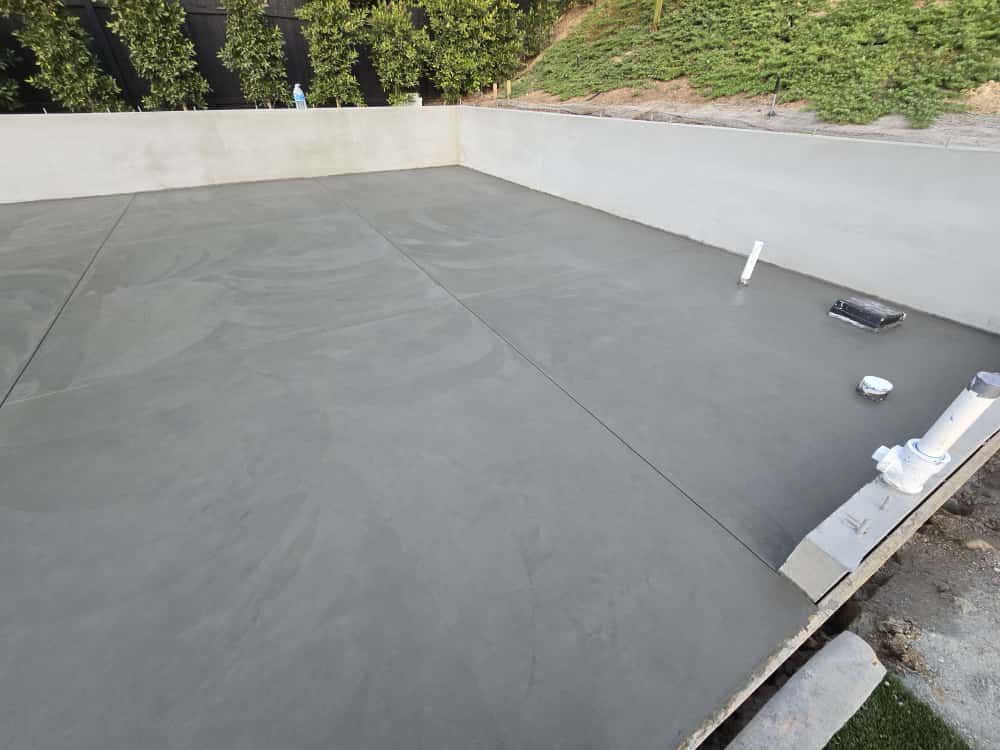 Flat gray rooftop with low parapet walls, vents, and construction materials near a wooded hillside.