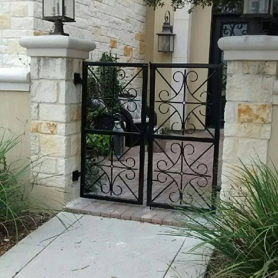 Black wrought-iron gate between stone pillars at a house entrance, with a concrete walkway and plants.