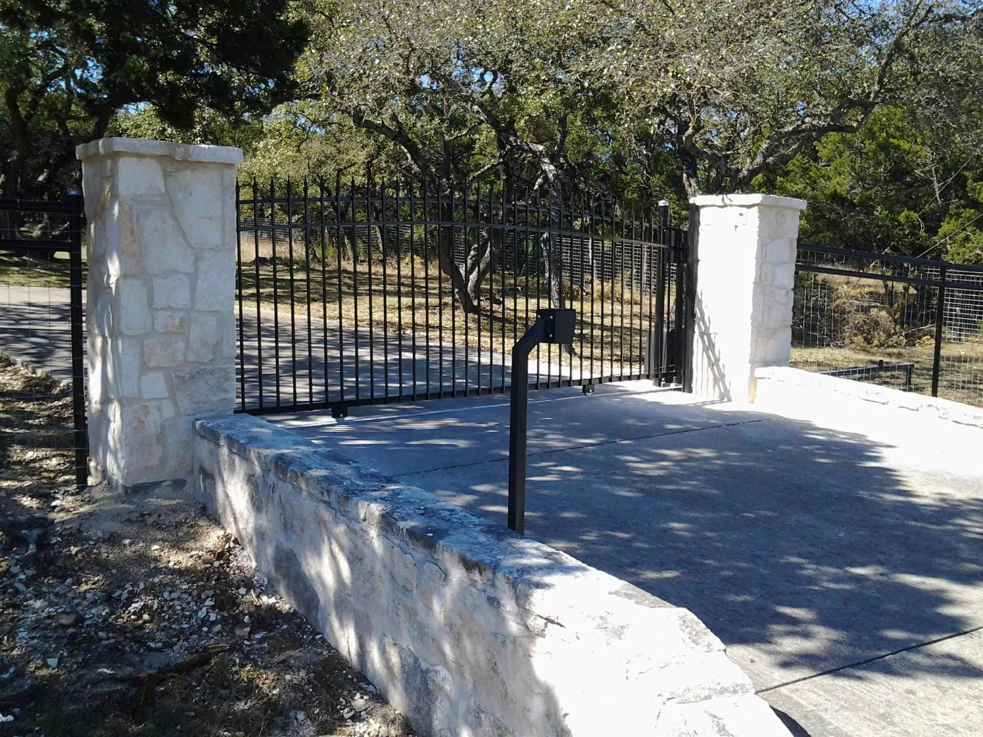Stone platform with a black handrail leading to a gated entrance between two pillars in a sunny park.
