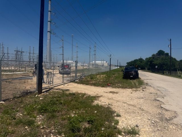 Gravel roadside beside a fenced electrical substation, with a black car parked under a clear blue sky.