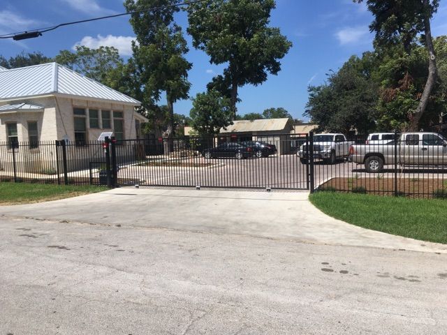 Gated driveway entrance to a small residential building with parked cars and trees under a blue sky