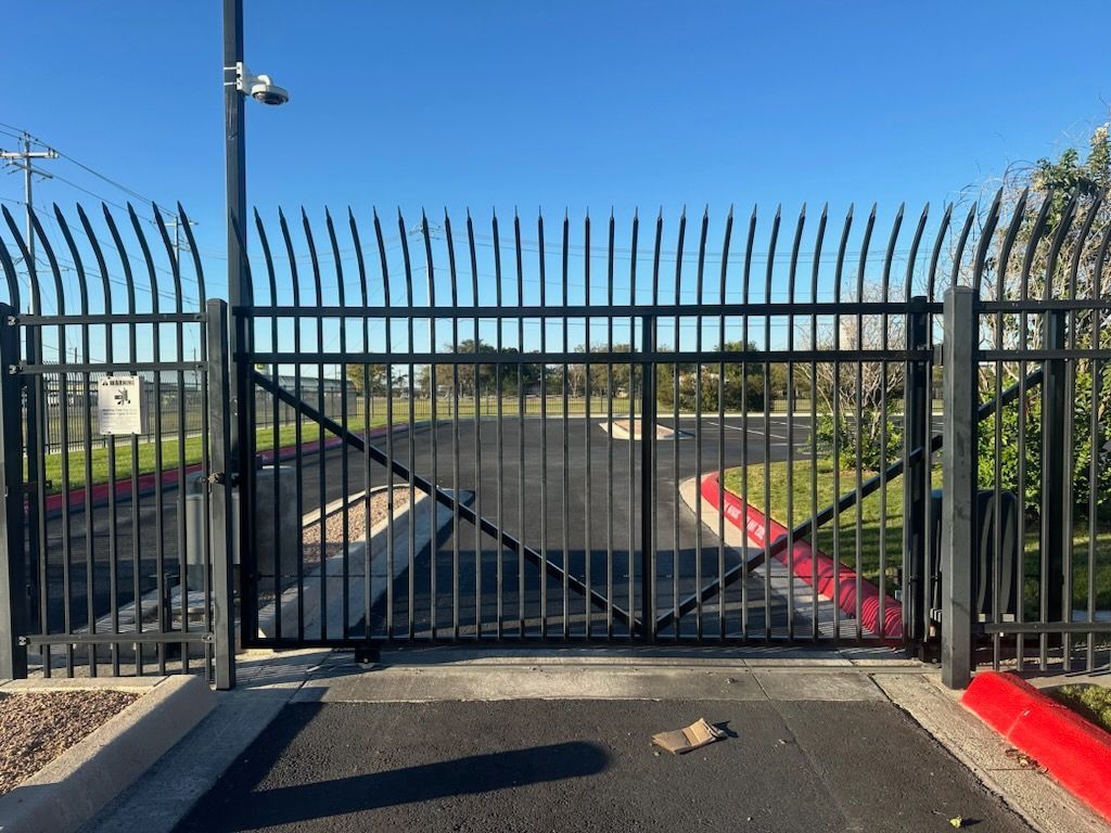 Closed black metal gate at a paved entrance, with red-and-white curbs and a clear blue sky beyond.