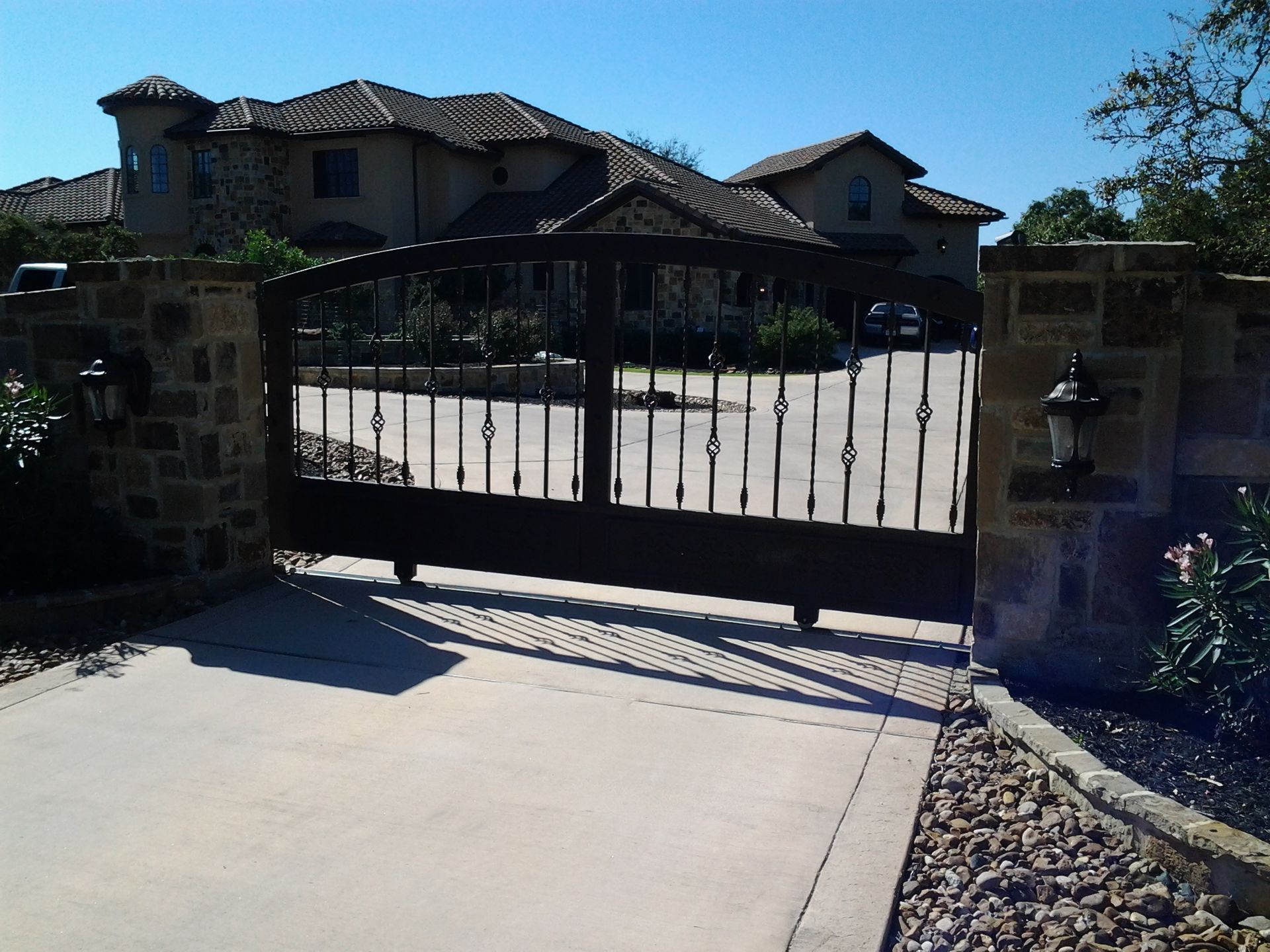 Black metal driveway gate in front of a house with stone pillars and tiled roof