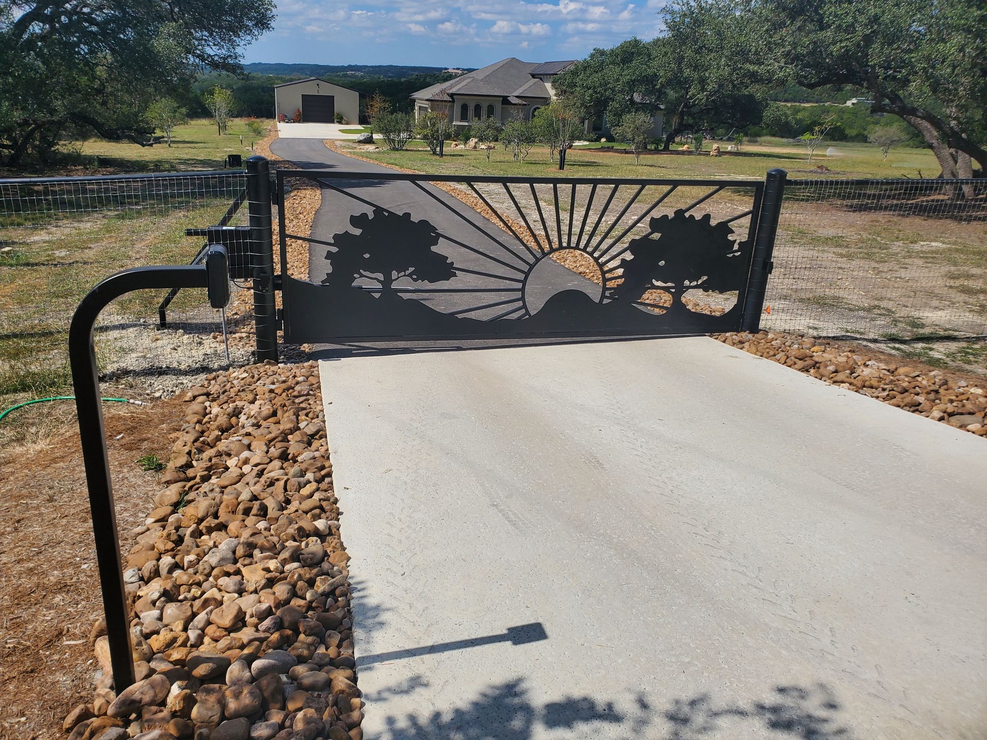 Black metal gate with sunburst design blocking a driveway in a rural yard