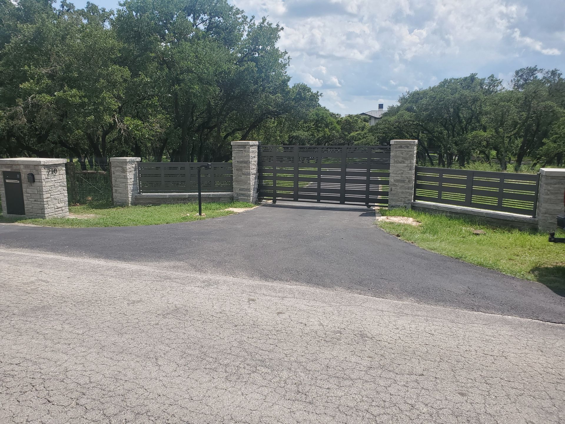Gravel driveway leading to a gated entrance with stone pillars and wooden fencing, surrounded by trees.