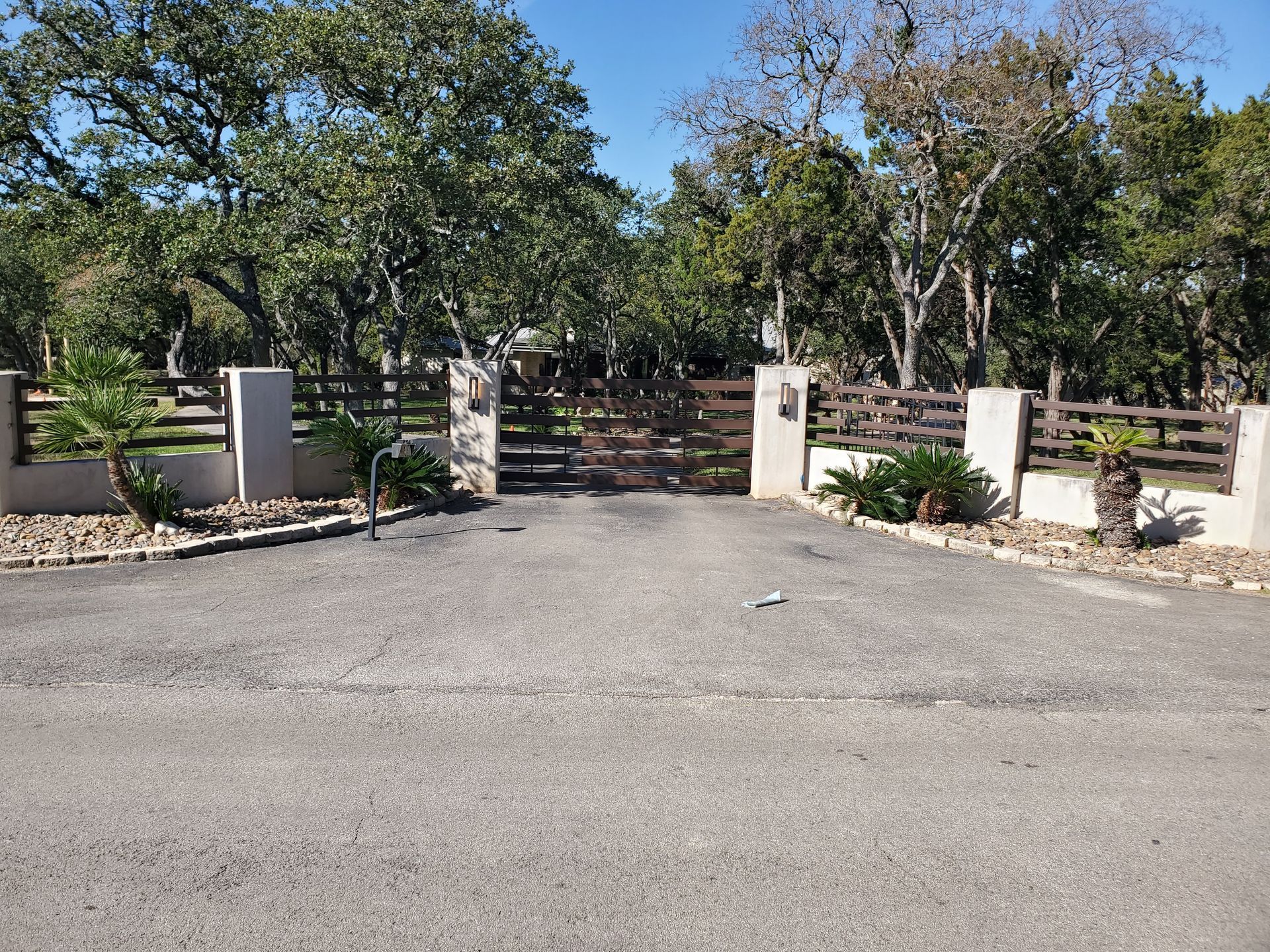 Gravel driveway entrance with stone pillars, wooden gate, and trees under a bright sky