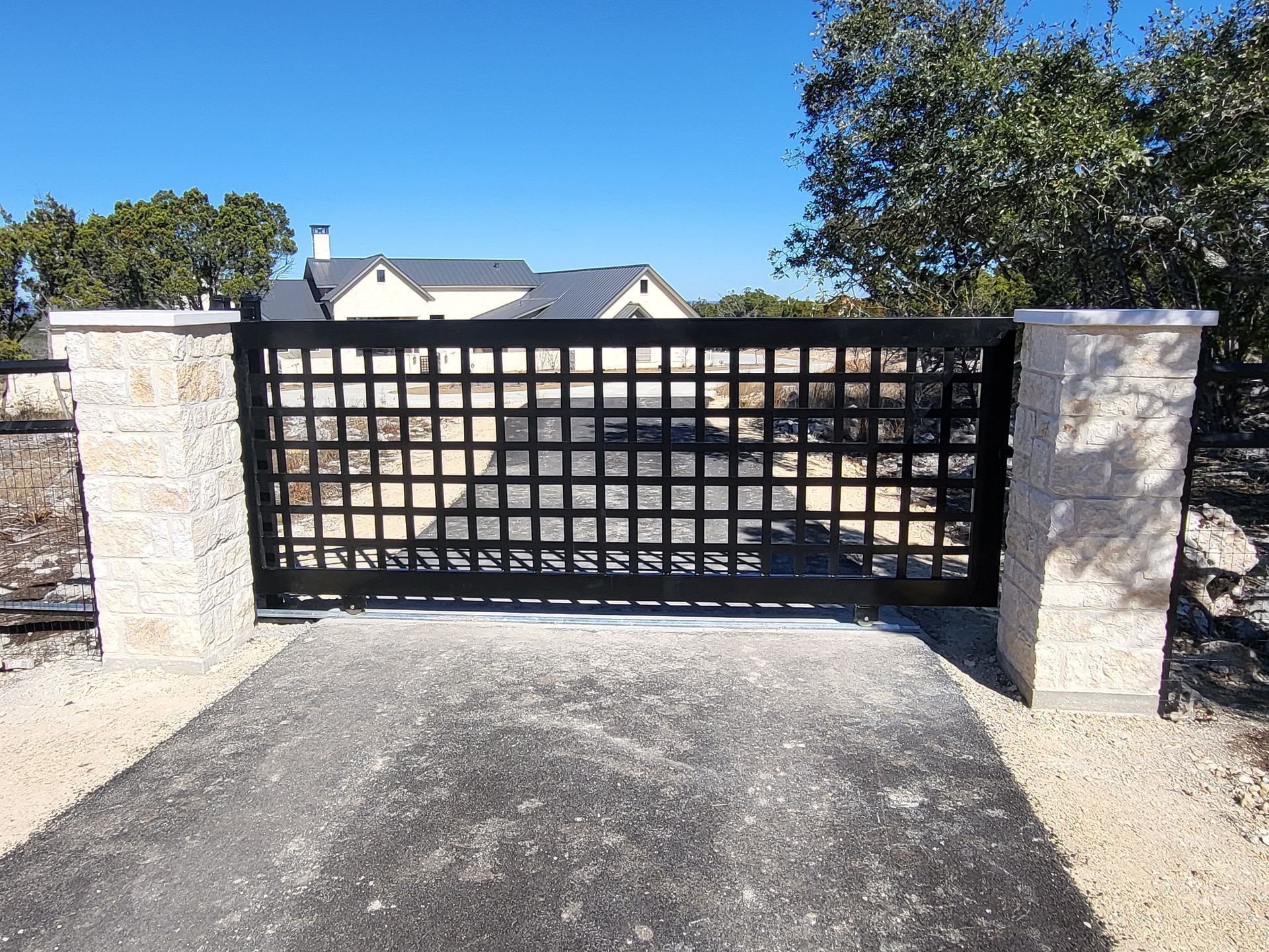 Black lattice driveway gate between stone pillars at the end of a gravel driveway under a clear blue sky