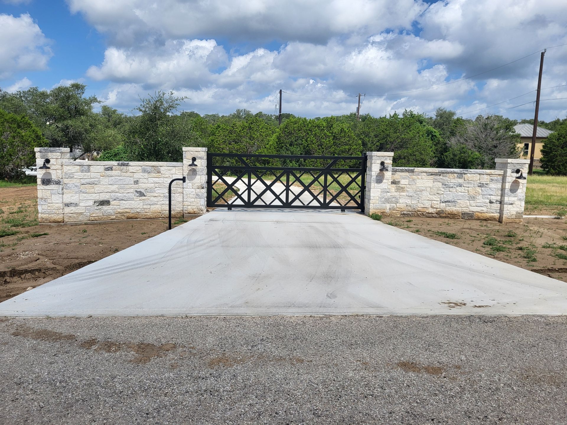 Concrete driveway leading to a closed black gate between white stone walls under a cloudy sky