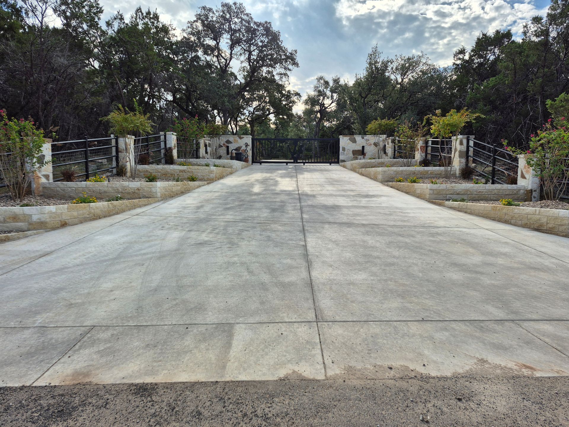 Wide concrete driveway leading to a closed black gate, flanked by stone walls and trees.