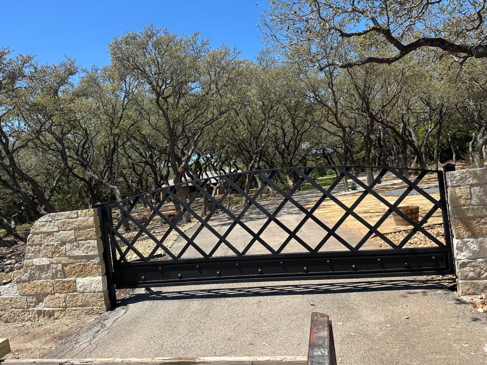 Black metal gate across a driveway, with stone wall and trees behind it.