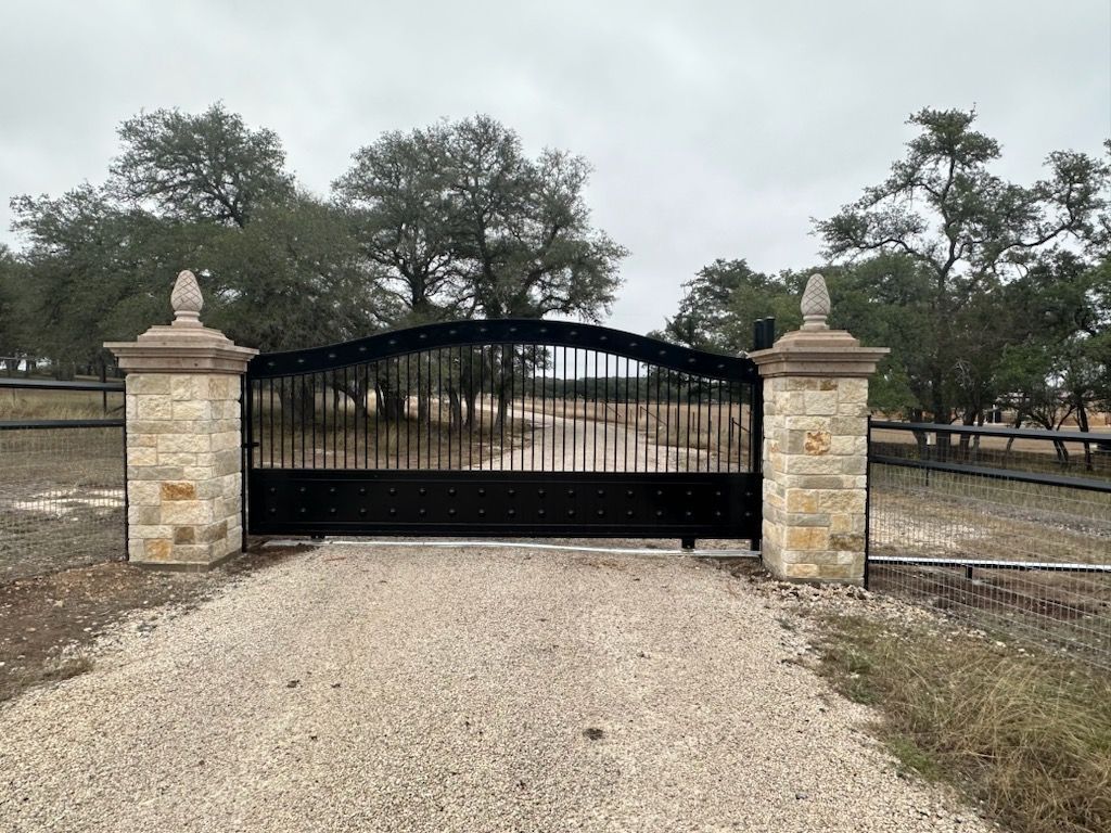 Black iron gate between stone pillars at the end of a gravel driveway, with trees beyond.