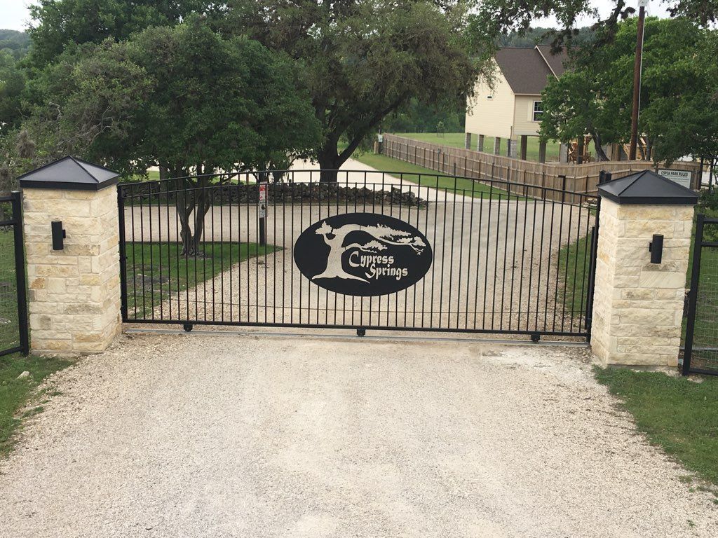 Closed black wrought-iron gate with stone posts at a driveway entrance, trees and a house beyond.