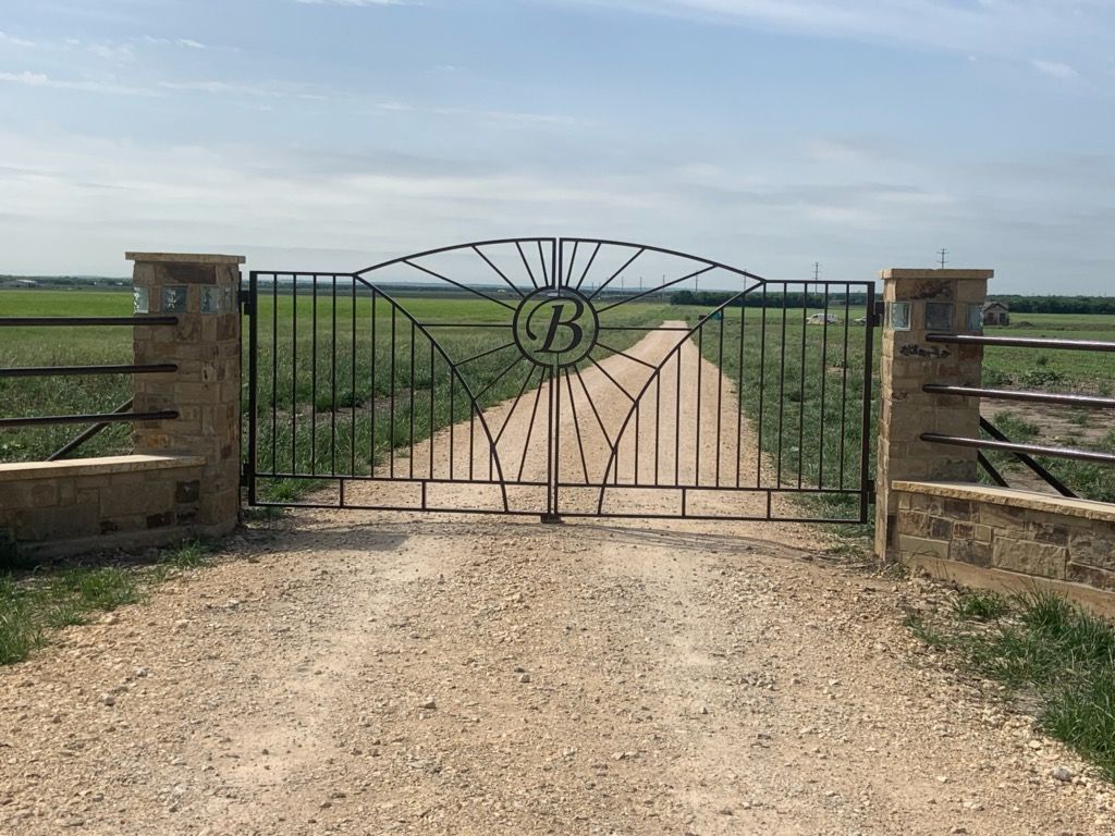 Rural driveway gate with a decorative monogram, flanked by stone pillars and open fields beyond