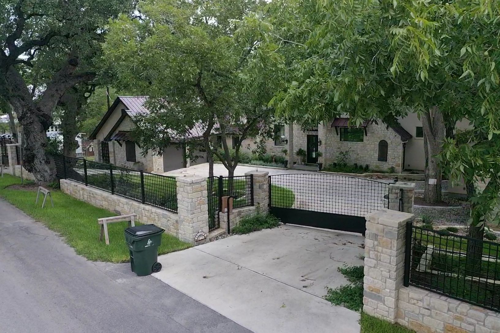Tree-lined residential driveway with stone walls, green gate, and a trash bin beside the street.