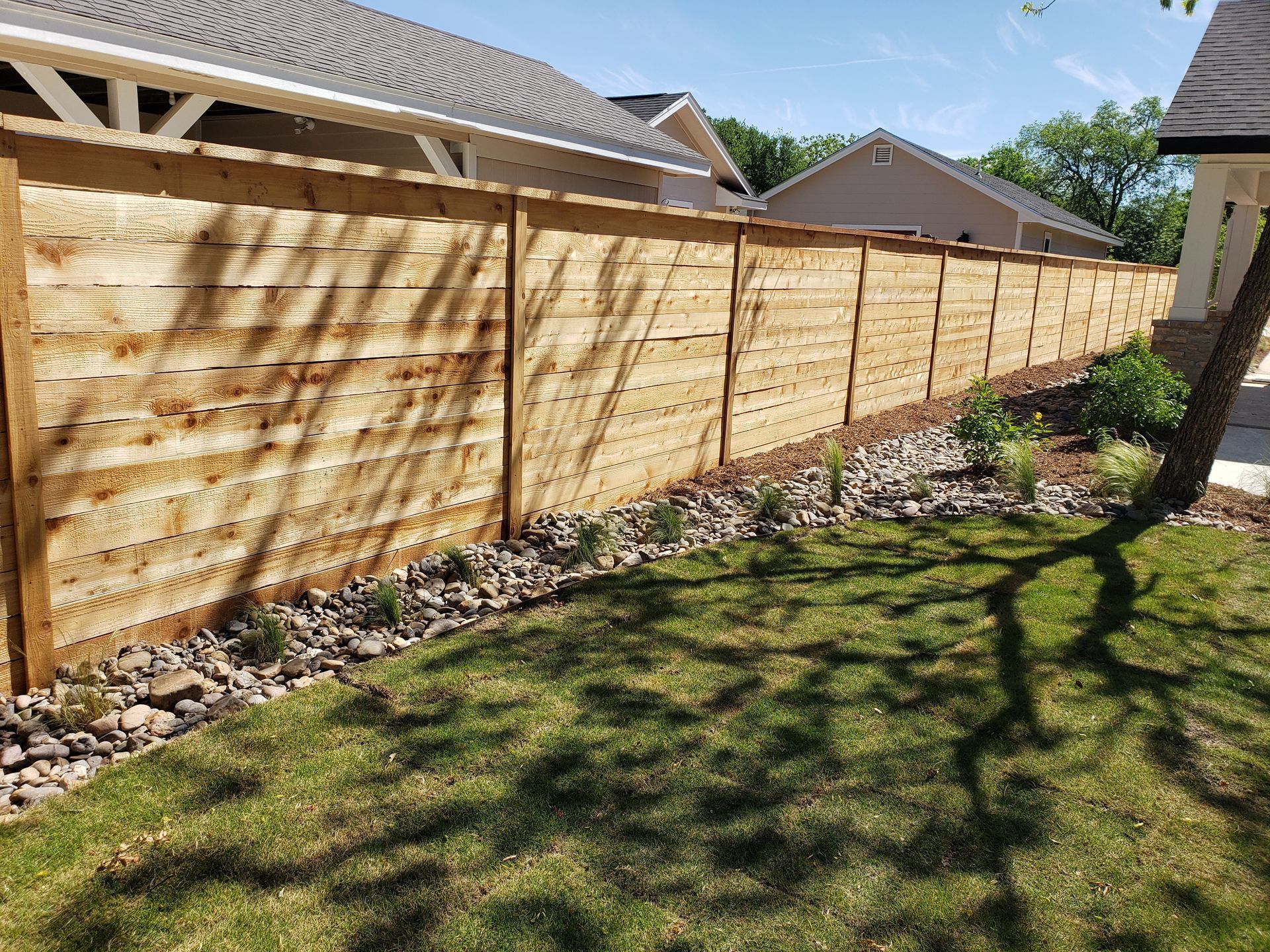 Wooden privacy fence lining a landscaped backyard with grass, rocks, and house roofs in the background