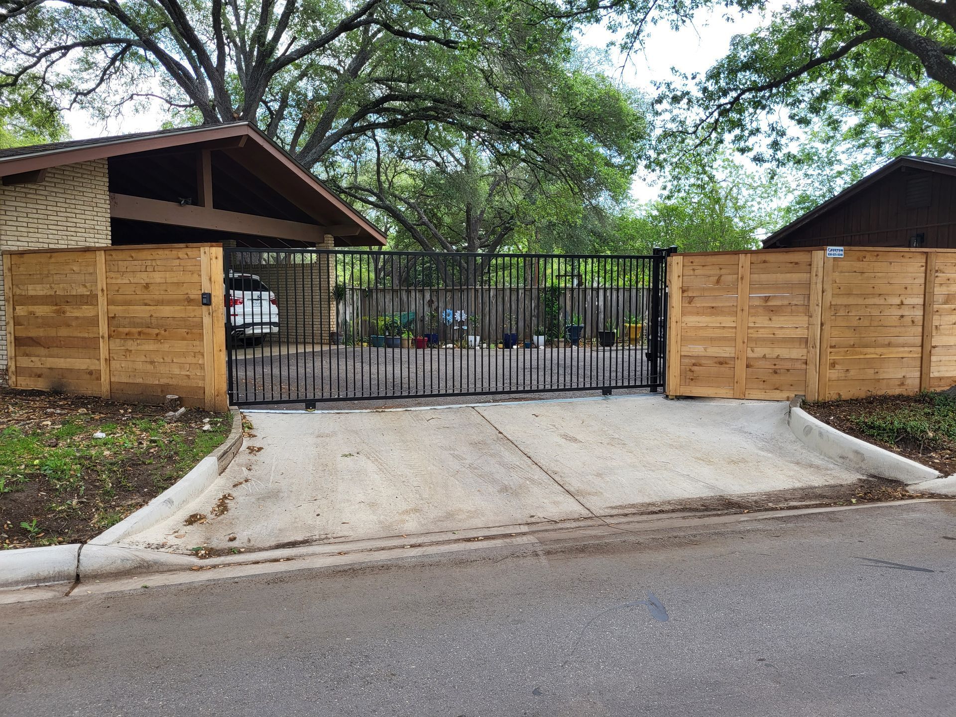 Wooden privacy fence and black gate at a driveway entrance, with trees and houses behind it.