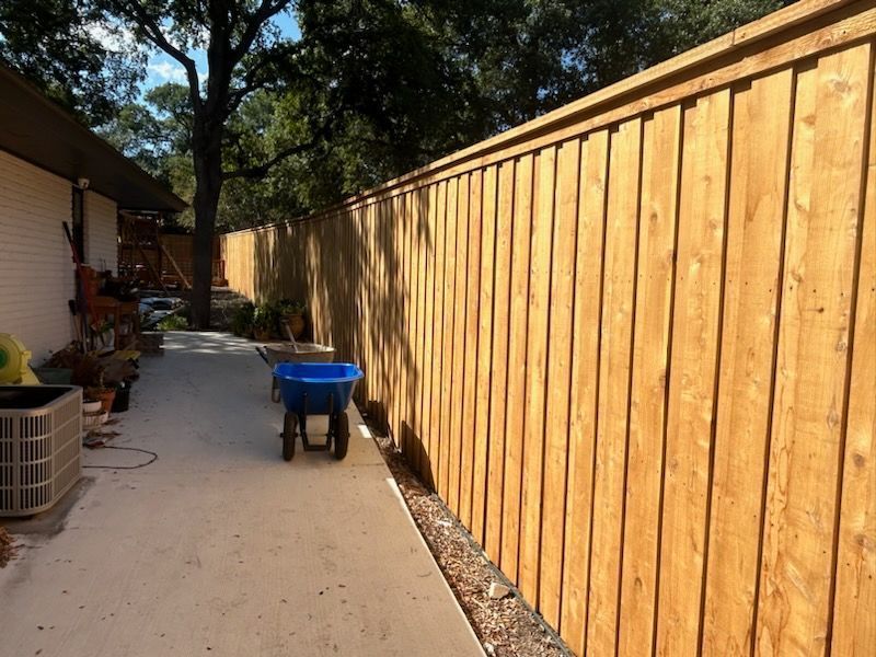 Narrow outdoor side yard with a long wooden fence, concrete path, and a blue wheelbarrow.