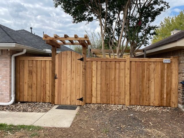 Wooden driveway gate and fence beside a brick house, with a small pergola over the gate.