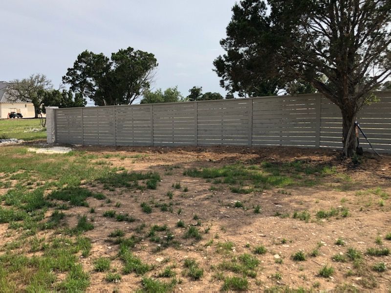 Sparse yard with patchy grass and a gray fence, with trees and a house in the background.
